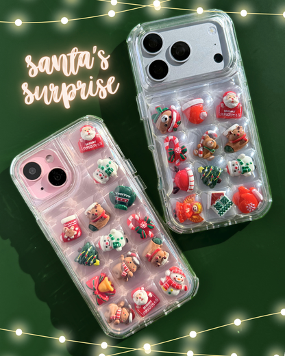 Two clear phone cases decorated with small Christmas-themed charms, shown on a dark green background with string lights and the text 'Santa's surprise'.