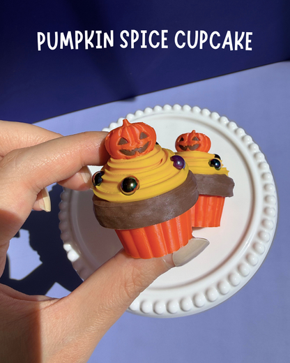 A hand holds a small pumpkin spice cupcake with orange frosting, yellow swirls, chocolate drizzle, and a jack-o'-lantern topper, with two more cupcakes on a white plate in the background. The text 'PUMPKIN SPICE CUPCAKE' appears at the top.